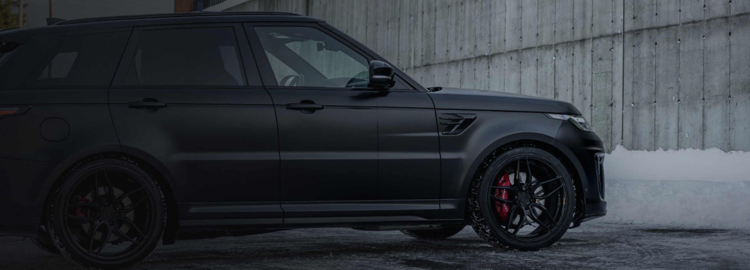 Matt svart Range Rover Sport med røde bremsekalipere foran en betongvegg i snøen